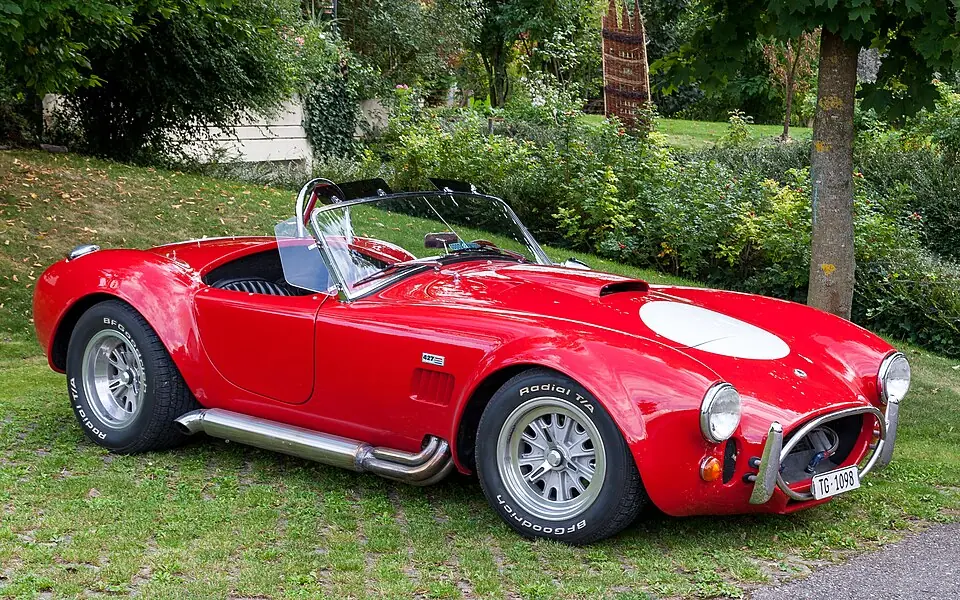 Red Shelby Cobra 427 roadster with white roundel, front three-quarter view, garden setting